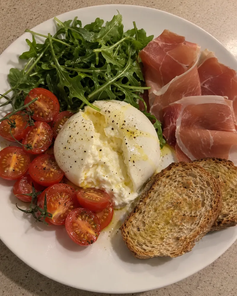 Burrata abierta con tomates cherry, jamón serrano, rúcula y pan integral tostado